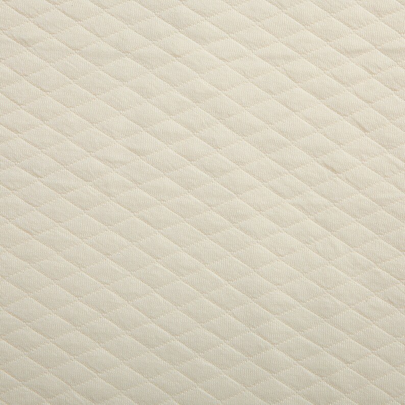 May include: Close-up of a cream-colored quilted fabric. The fabric has a repeating diamond pattern, creating a textured surface. The material appears soft and is likely used for bedding or upholstery.