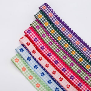 May include: Assortment of colorful gingham ribbon trim with scalloped edges and embroidered floral designs. The ribbons are in shades of green, purple, pink, blue, and white.