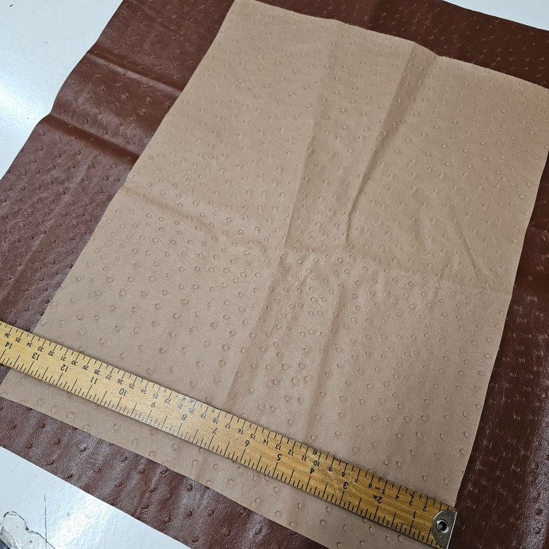 May include: A close-up of two pieces of leather, one brown and one beige. The beige leather has a textured surface that resembles ostrich skin. A wooden ruler is placed on top of the beige leather.