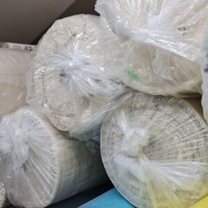Puede incluir: Primer plano de varios rollos de tela envueltos en pl&aacute;stico transparente. La tela es de color beige claro y est&aacute; enrollada con fuerza. Los rollos est&aacute;n apilados uno encima del otro.