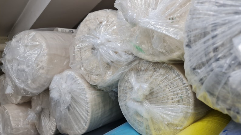 Puede incluir: Primer plano de varios rollos de tela envueltos en pl&aacute;stico transparente. La tela es de color beige claro y est&aacute; enrollada con fuerza. Los rollos est&aacute;n apilados uno encima del otro.