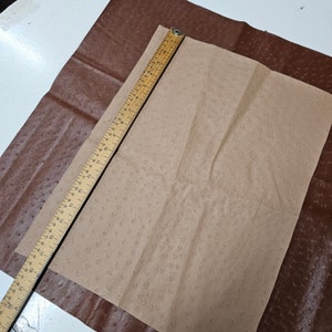 May include: A brown leather sheet with a smaller sheet of beige leather with a textured pattern on top. A wooden ruler is placed on top of the beige sheet.