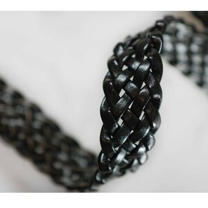 Peut inclure: Gros plan sur une sangle tress&eacute;e en cuir noir. La sangle est tiss&eacute;e en un motif serr&eacute; et complexe.