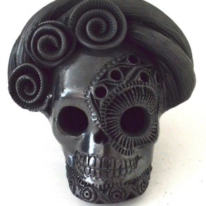 Puede incluir: Cráneo de cerámica negro con detalles intrincados, incluyendo un diseño floral en la frente y un ojo decorativo. El cráneo es una decoración tradicional del Día de Muertos.