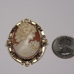May include: A gold-tone cameo brooch with a carved shell cameo of a woman's profile. The cameo is set in a gold-tone frame with ornate details. The brooch is about the size of a quarter.