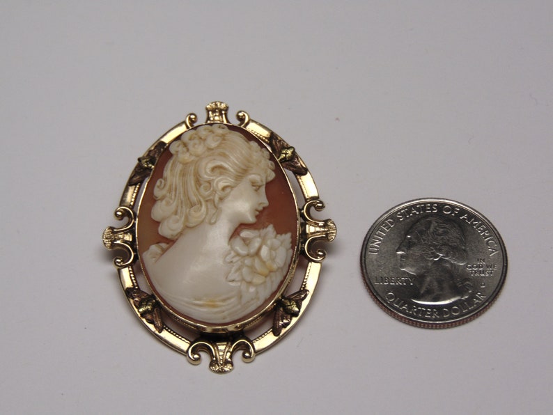 May include: A gold-tone cameo brooch with a carved shell cameo of a woman's profile. The cameo is set in a gold-tone frame with ornate details. The brooch is about the size of a quarter.