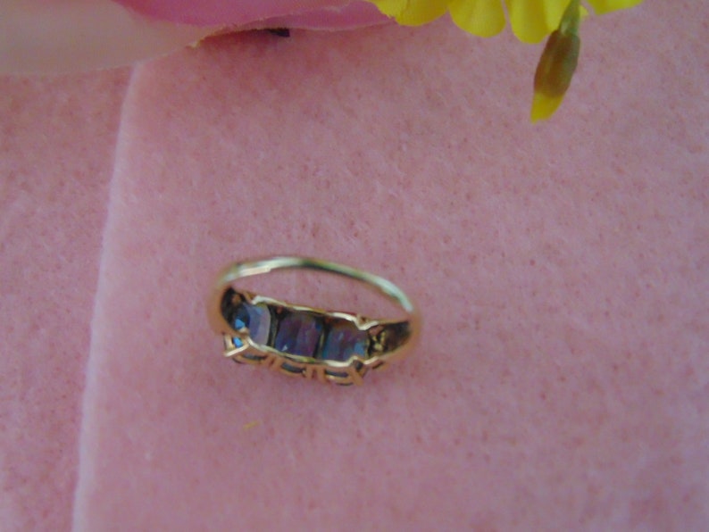 Pode incluir: Um anel de ouro com cinco pedras preciosas azuis engastadas em fila. O anel est&aacute; sobre uma superf&iacute;cie rosa.