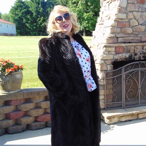 May include: A long, black fur coat with a shawl collar. The coat is worn by a person with blonde hair and sunglasses. The person is standing in front of a stone fireplace.