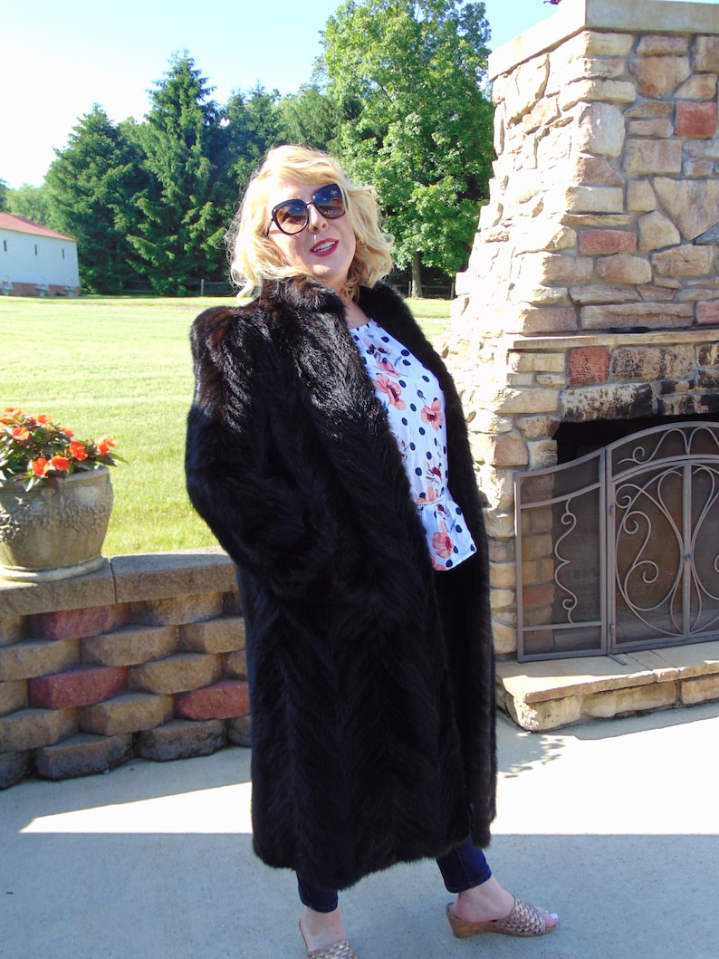 May include: A long, black fur coat with a shawl collar. The coat is worn by a person with blonde hair and sunglasses. The person is standing in front of a stone fireplace.