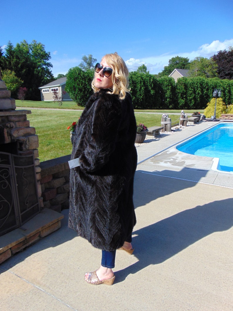 May include: A woman wearing a long, black fur coat and sunglasses stands near a swimming pool. She is wearing blue jeans and brown sandals.