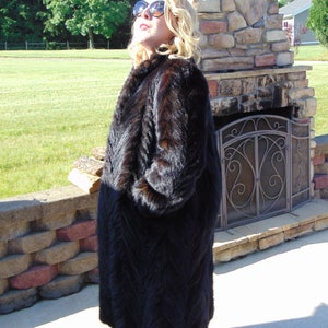May include: A long, black fur coat with a chevron pattern. The coat is worn by a person standing in front of a stone fireplace.