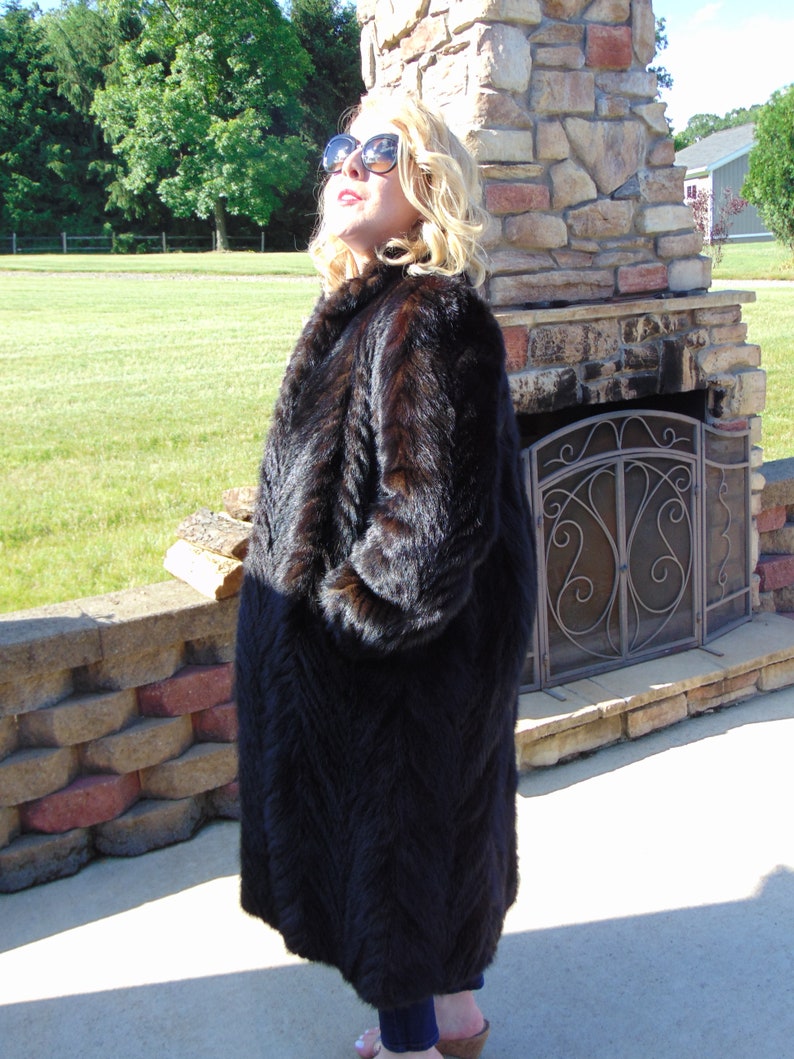 May include: A long, black fur coat with a chevron pattern. The coat is worn by a person standing in front of a stone fireplace.