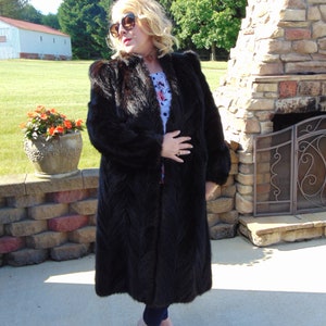 May include: A long black fur coat with a chevron pattern. The coat is worn by a person standing in front of a stone fireplace.