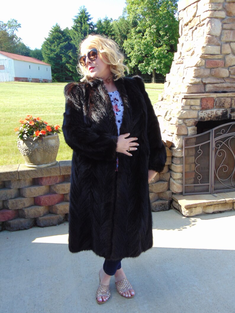 May include: A long black fur coat with a chevron pattern. The coat is worn by a person standing in front of a stone fireplace.