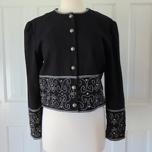 May include: A black cropped jacket with silver buttons and a silver embroidered trim around the bottom and cuffs.
