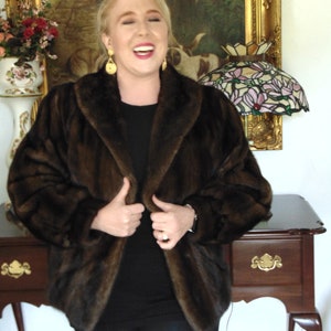 May include: A woman wearing a brown fur jacket with a black top and black pants. She is smiling and holding the jacket closed. She is standing in front of a wooden table with a gold-framed picture and a stained glass lamp.