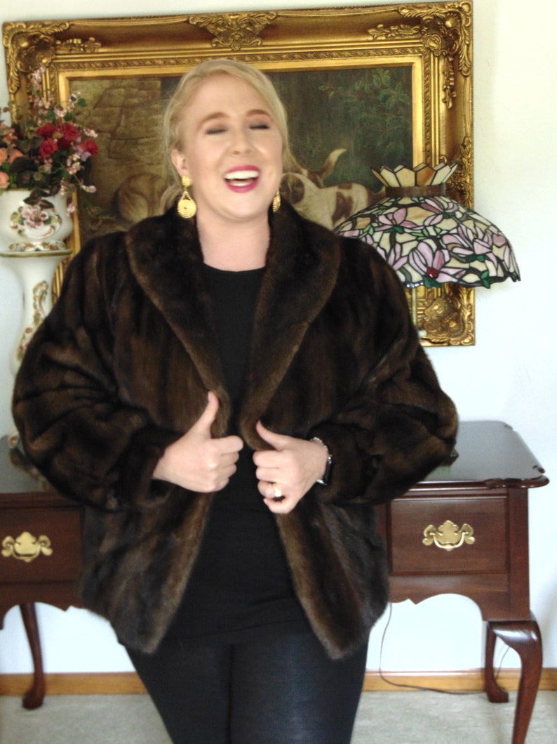 May include: A woman wearing a brown fur jacket with a black top and black pants. She is smiling and holding the jacket closed. She is standing in front of a wooden table with a gold-framed picture and a stained glass lamp.