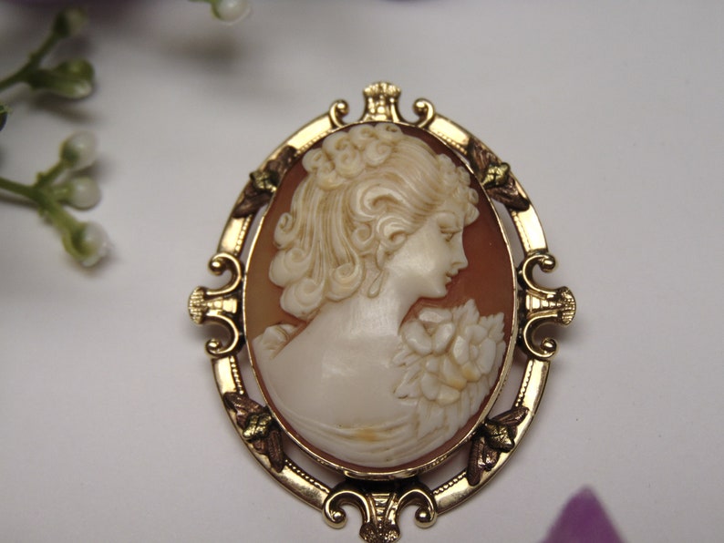 May include: A gold-toned cameo brooch featuring a woman's profile with a floral design. The cameo is set in a detailed oval frame.
