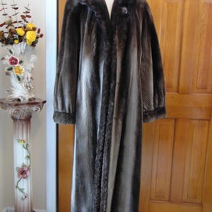 May include: A long, brown fur coat with a shawl collar and a single button closure. The coat is made of a soft, luxurious fur and has a sleek, elegant design.