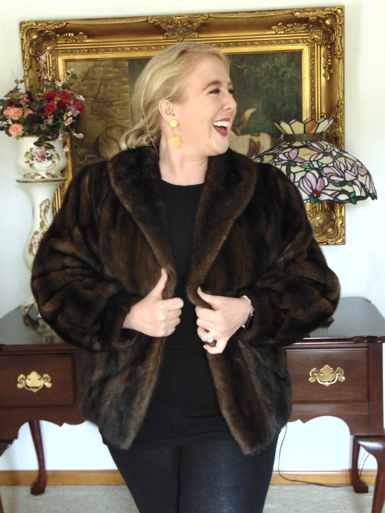 May include: A woman wearing a brown fur coat with a black shirt and black pants. She is standing in front of a gold framed picture with a painting inside.