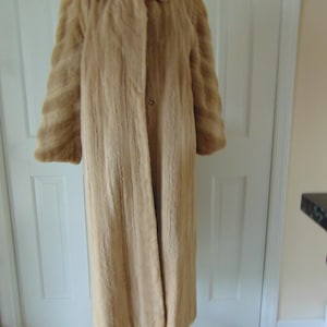 May include: A long, light brown fur coat with a notched collar and long sleeves. The coat is buttoned at the waist and hangs to the ankles. The coat is displayed on a wooden dress form.