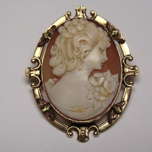 May include: A gold-tone cameo brooch featuring a carved shell cameo of a woman with a flower. The cameo is set in a detailed oval frame with gold accents.