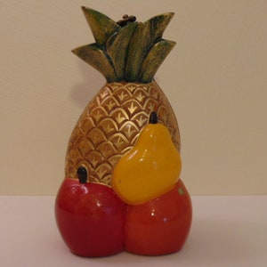 Può includere: Una scultura di frutta in ceramica con un ananas, una pera e due mele. L'ananas è dorata con la cima verde, la pera è gialla e le mele sono rosse.