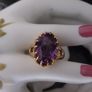 May include: A gold ring featuring a large, oval-cut purple gemstone. The ring has an ornate, scalloped setting and is displayed on a white surface. The gemstone's color is a deep, rich purple.