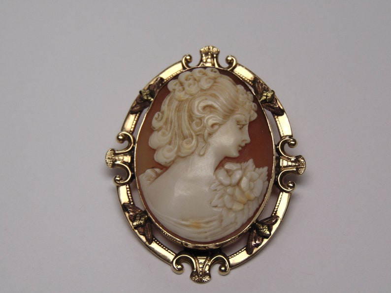 May include: A gold-tone cameo brooch with a carved shell cameo of a woman's profile. The cameo is set in a gold-tone frame with ornate details and green accents.
