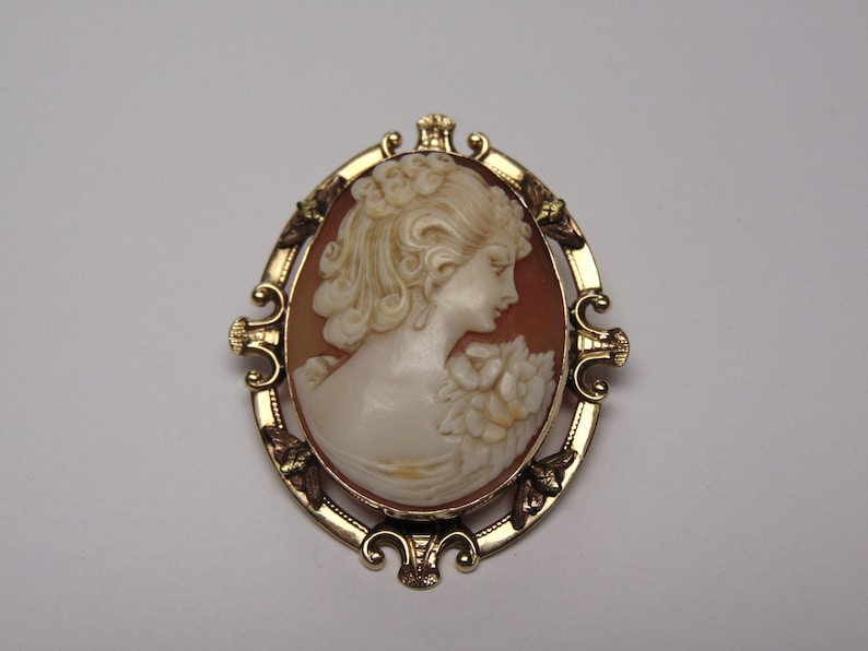 May include: A gold-toned cameo brooch featuring a carved shell cameo of a woman's profile with a floral design. The cameo is set in a gold-toned oval frame with ornate details.