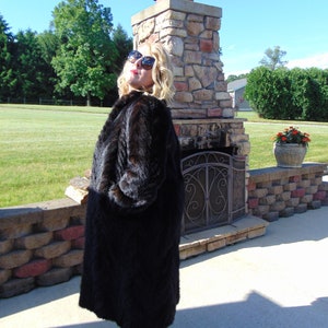 May include: A woman wearing a long black fur coat and sunglasses stands in front of a stone fireplace. She is wearing a pair of gold sandals.