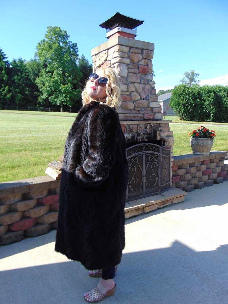 May include: A woman wearing a long black fur coat and sunglasses stands in front of a stone fireplace. She is wearing a pair of gold sandals.