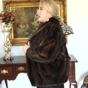 May include: A brown fur jacket with a long sleeve and a collar. The jacket is worn by a person with blonde hair and is standing in front of a gold framed picture.