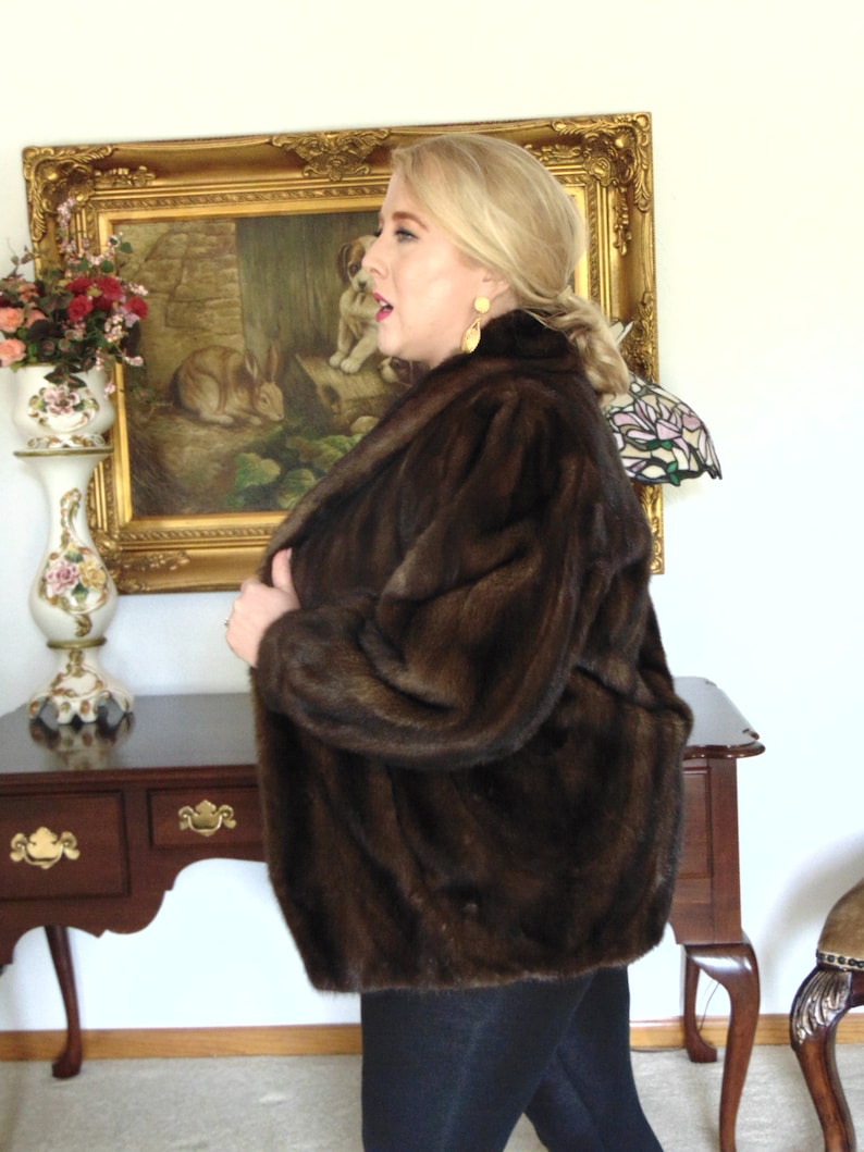 May include: A brown fur jacket with a long sleeve and a collar. The jacket is worn by a person with blonde hair and is standing in front of a gold framed picture.