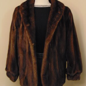 May include: A brown fur jacket with a large collar and long sleeves. The jacket is on a mannequin and is displayed against a light beige wall.