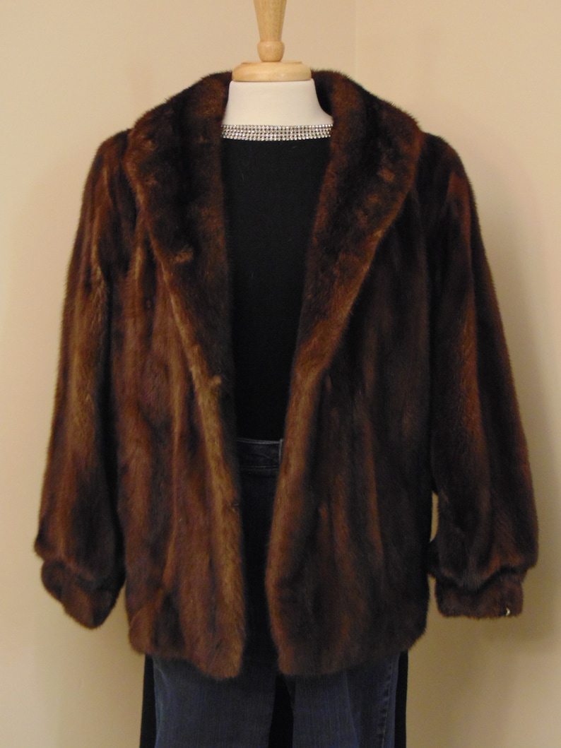 May include: A brown fur jacket with a large collar and long sleeves. The jacket is on a mannequin and is displayed against a light beige wall.