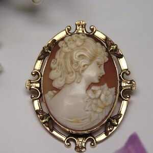 May include: A gold-toned cameo brooch with a carved shell cameo of a woman's profile. The cameo is set in a gold-toned frame with ornate details and green accents.