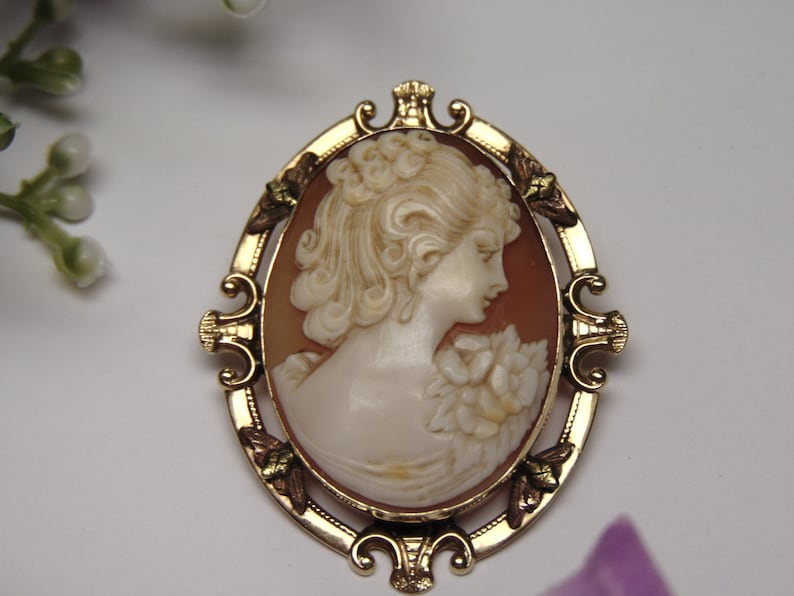 May include: A gold-toned cameo brooch with a carved shell cameo of a woman's profile. The cameo is set in a gold-toned frame with ornate details and green accents.