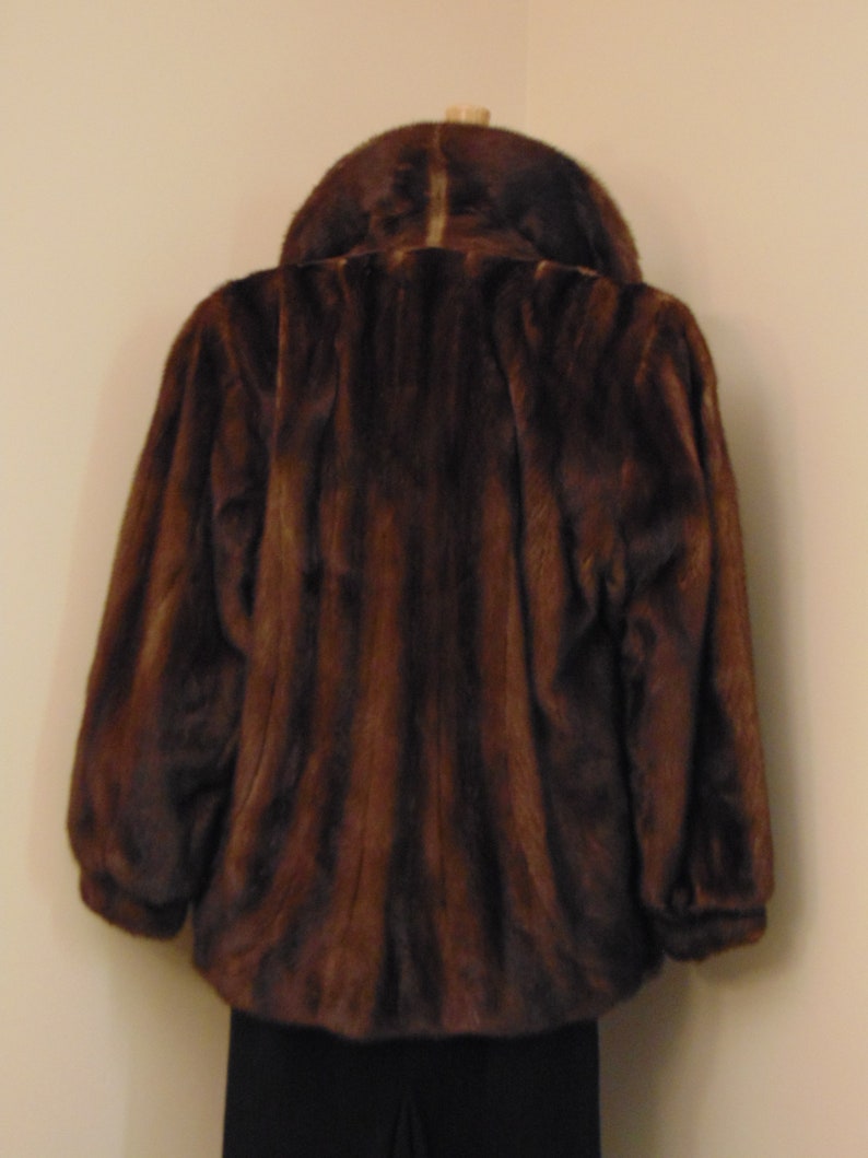 May include: A brown fur jacket with a collar. The jacket is on a mannequin and the background is a light beige wall.
