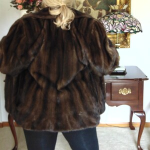 May include: A brown fur coat with a long sleeve design. The coat is worn by a person with long blonde hair. The person is standing in front of a wall with a painting and a lamp.