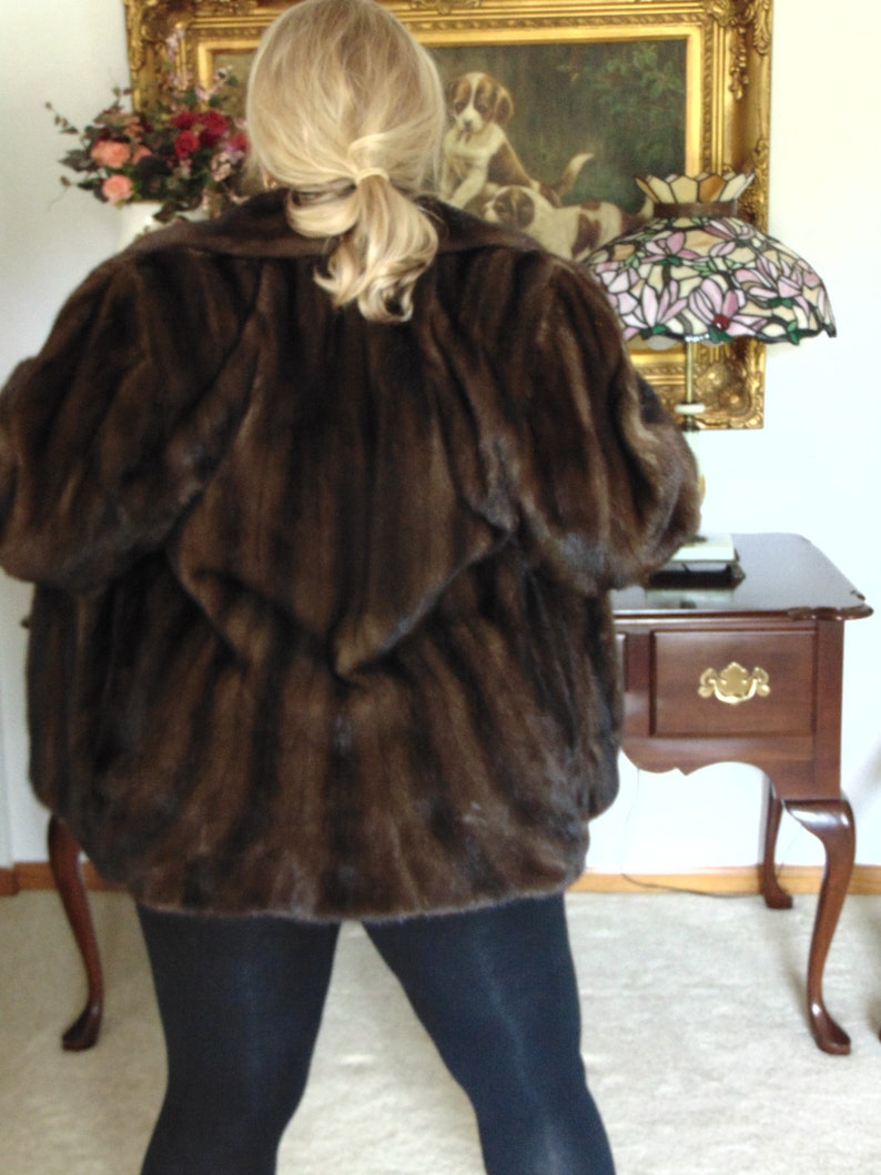 May include: A brown fur coat with a long sleeve design. The coat is worn by a person with long blonde hair. The person is standing in front of a wall with a painting and a lamp.