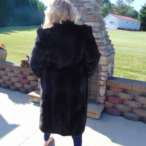 May include: A long black fur coat with a stand-up collar. The coat is worn by a person standing in front of a stone wall.