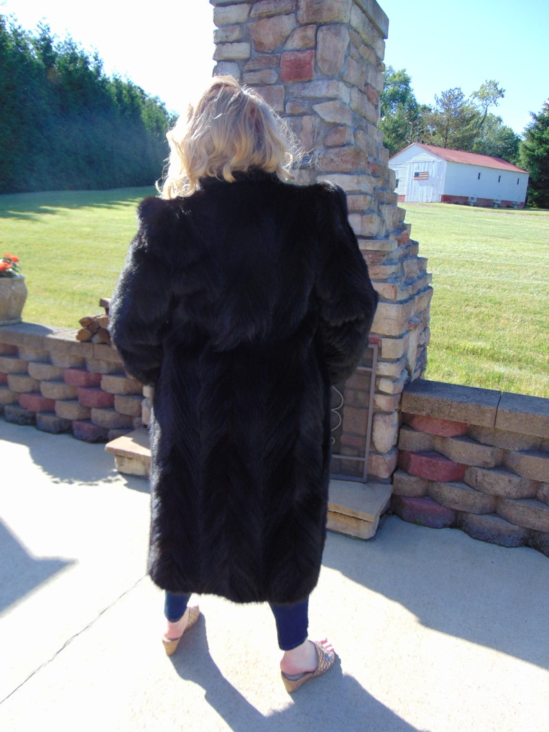May include: A long black fur coat with a stand-up collar. The coat is worn by a person standing in front of a stone wall.