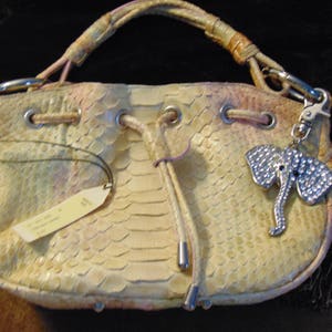 Könnte beinhalten: Eine kleine, helle Handtasche aus Schlangenleder mit einem silbernen Elefanten-Anhänger. Die Tasche hat einen Kordelverschluss und eine rosa-weiße Farbgebung.