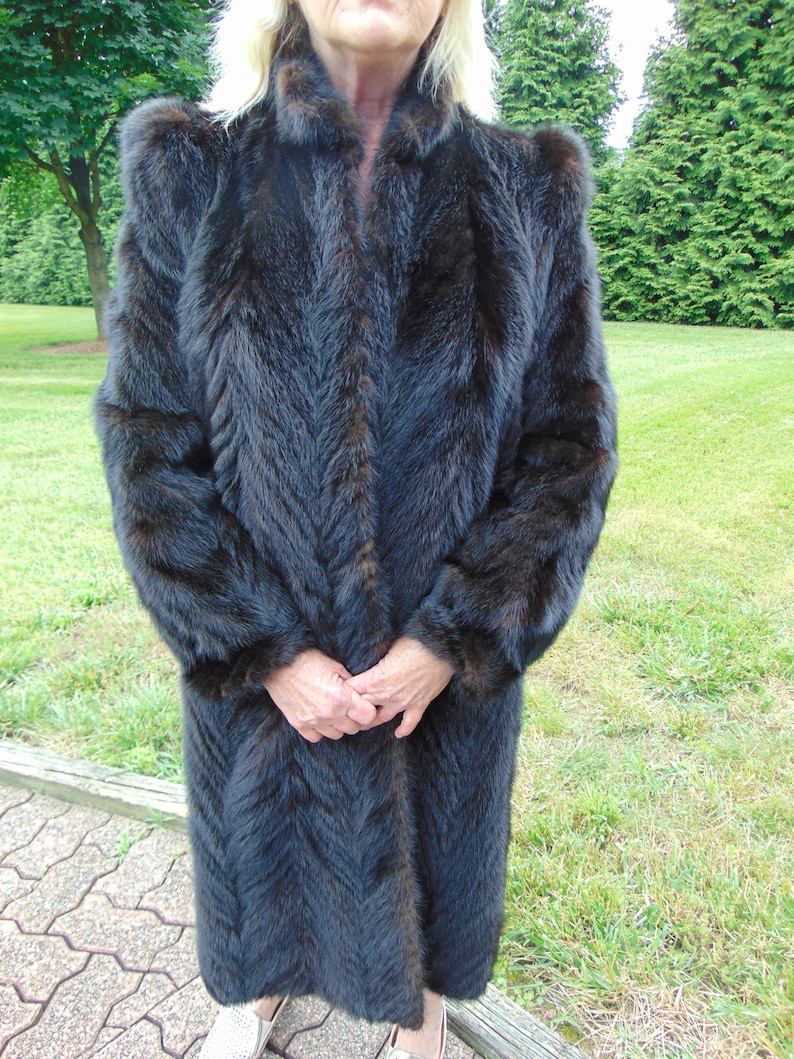 May include: A long, dark brown fur coat with a chevron pattern. The coat has a high collar and long sleeves.