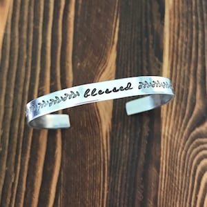 May include: A silver-colored cuff bracelet with the word "blessed" in a script font. The bracelet has a leaf design on each end. The background is a dark brown wooden surface.