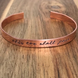 May include: A copper bracelet with the words "this too shall pass" engraved on it.