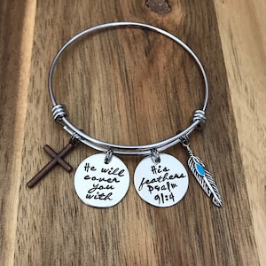 May include: A silver expandable bracelet with three charms. The charms are a cross, a round charm with the text "He will cover you with" and a round charm with the text "His feathers Psalm 91:4". The bracelet also has a small feather charm with a blue accent.