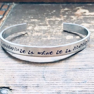 May include: Silver metal bracelet with the inscription "it is what it is" and a leafy design on either side.