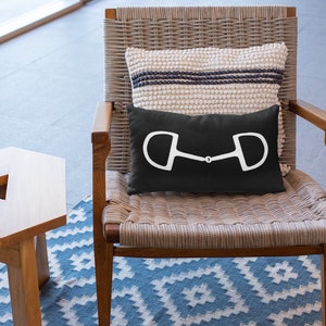 May include: A black pillow with a white horse bit design sits on a wicker chair with a white and gray striped pillow behind it. The chair is on a blue and white patterned rug.